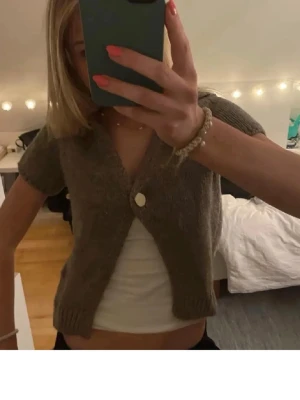 zara kofta - Kortärmad brun kofta i mjukt stickat material med en stor vit knapp framtill. Modellen är croppad och har ribbade kanter vid ärmslut och nederkant. Perfekt att slänga över ett linne för en chill look.