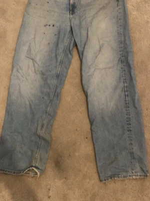 Ljusblå raka jeans - Ett par ljusblå jeans med rak passform och klassisk femficksdesign. Jeansen har slitna detaljer vid bensluten och några mörka fläckar framtill. Tillverkade i denim med en avslappnad look, perfekta för en casual stil.