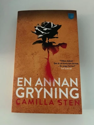 En annan gryning av Camilla Sten - En dystopisk thrillerbok med orange och svart omslag, prydd med en svartvit ros och blodröd detalj. Boken är skriven av Camilla Sten och utgiven av Pocketförlaget. Perfekt för dig som gillar spänning och samhällskritik i romanform.
