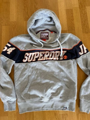 Grå Superdry hoodie med tryck - Snygg grå hoodie från Superdry med stort tryck över bröstet och svarta detaljer. Har huva med dragsko, känguruficka och sportiga siffror på ärmarna. Perfekt för en avslappnad streetstyle-look.