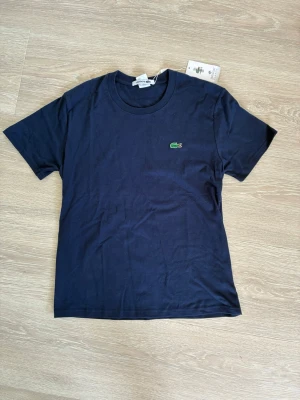 Marinblå t-shirt från Lacoste - Snygg marinblå t-shirt från Lacoste med klassisk rund halsringning och den ikoniska gröna krokodilloggan broderad på bröstet. Tillverkad i mjuk bomull och har en enkel, stilren design som passar till allt.