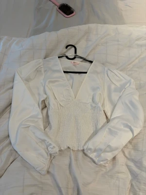 Vit blus med puffärmar - Snygg vit blus med djup v-ringning, puffiga långärmar och smockad midja som ger en feminin siluett. Tillverkad i ett glansigt, satinliknande material som känns lyxigt och trendigt. Perfekt att styla med jeans eller kjol för en chic look. Passar som S och M