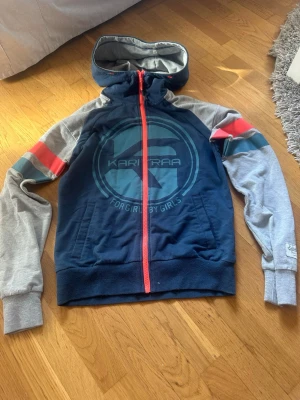 Blå och grå hoodie Kari Traa - Sportig hoodie från Kari Traa i marinblått med grå ärmar. Stora loggan framtill, röda och turkosa ränder på ärmarna samt röd dragkedja. Mjuk huva och ribbade muddar. Perfekt för all träning, samt väldigt luftig. Har själv använt den några få gånger när jag har tränat. Den är i storlek S.