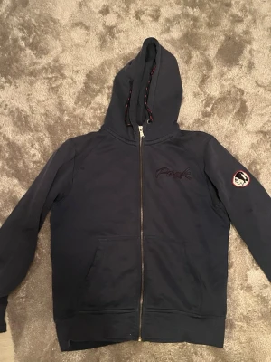 Mörkblå hoodie med dragkedja - Snygg mörkblå hoodie med dragkedja och broderad Peak-logga på bröstet. Huva med dragsko och två fickor framtill. Patch med motiv på ena ärmen. Perfekt för en avslappnad stil.