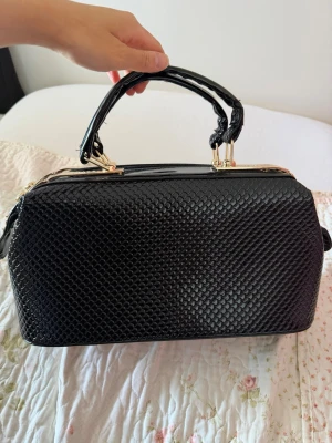 Svart handväska med ormmönster - Snygg svart handväska i skinnande konstläder med ormmönstrad struktur. Väskan har guldfärgade detaljer och dubbla handtag som är tvinnade för en cool look. Perfekt storlek och stängs med dragkedja upptill.
