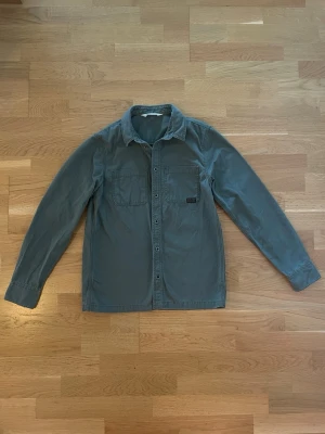 Grön overshirt från H&M - Snygg grön overshirt från H&M med två bröstfickor och knappar framtill. Skjortan har en klassisk krage och är tillverkad i bomull, perfekt att bära som lager över en t-shirt. Enkel och stilren design som passar till mycket.