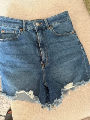 Blå högmidjade jeansshorts - Säljer ett par blå jeansshorts med hög midja och råa, fransiga kanter. Klassisk femficksmodell med dragkedja och knapp framtill. Perfekta för varma dagar och ger en avslappnad vibe.