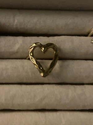 Guldig ring med hjärtformad design - Unik ring i guldfärgad metall med öppen hjärtformad design. Ringen har en organisk, lite vågig struktur som ger en trendig och modern känsla. Den är justerbar. 