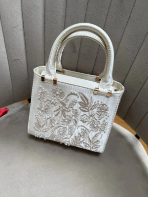 Vit handväska med broderade blommor - Snygg vit handväska i boxig modell med två handtag och guldfärgade detaljer. Väskan har vackra broderade blommor och blad på framsidan samt magnetlås upptill.