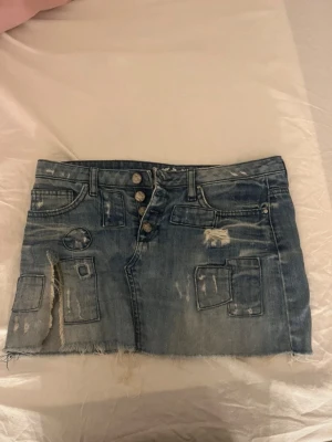 Kort jeanskjol med slitningar och lappar - Säljer en blå kort jeanskjol med slitna detaljer och flera dekorativa lappar. Kjolen har knappar framtill och fickor både fram och bak. Perfekt för dig som gillar en edgy och avslappnad stil.