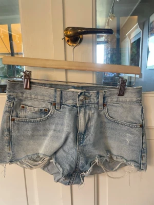 Ljusblå jeansshorts med fransar - Säljer ett par ljusblå denimshorts med råa, fransiga kanter och klassisk femficksdesign. Shortsen har normal passform och bälteshällor i midjan. Perfekta för varma dagar och ger en avslappnad look.