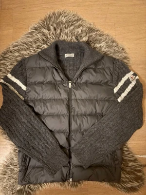 Grå Moncler cardigan med stickade ärmar  S - Snygg grå cardigan / jacka från Moncler med stickade ärmar i mörkgrått och vita ränder. Jackan har dragkedja framtill, hög krage och Moncler-logga på ärmen. Perfekt för lager-på-lager och streetstyle. Materialmix av dun och stickat ger en unik look. Perfekt till hösten som kommer så passa på! Aldrig använd, passar även XS skulle jag säga.