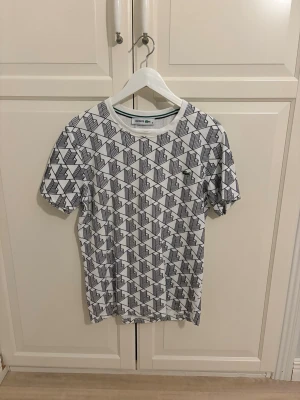 Vit mönstrad t-shirt från Lacoste - Säljer en vit t-shirt från Lacoste med svart geometriskt mönster över hela tröjan och den klassiska gröna krokodilloggan på bröstet. T-shirten har rund hals och korta ärmar. Perfekt för dig som gillar stilrena men ändå unika plagg.
