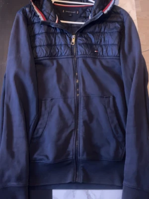 Marinblå vindjacka från Tommy Hilfiger - Snygg marinblå vindjacka från Tommy Hilfiger med dragkedja framtill och två stora fickor. Jackan har quiltade detaljer upptill och klassiska Hilfiger-ränder i kragen. Perfekt för dig som vill ha en stilren och sportig look. 1000-2000kr ny pris 