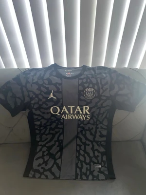 Svart PSG matchtröja med mönster - Snygg svart PSG matchtröja från Jordan med grått djurmönster och Qatar Airways-logga på bröstet. Tröjan har rund hals, korta ärmar och klubbmärke på vänster sida. Perfekt för dig som älskar fotboll och vill sticka ut på planen.