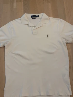 Beige pikétröja från Polo Ralph Lauren - Klassisk beige pikétröja från Polo Ralph Lauren med korta ärmar och krage. Tröjan har knappslå framtill och den ikoniska broderade loggan på bröstet. Perfekt till jeans eller shorts för en stilren look.