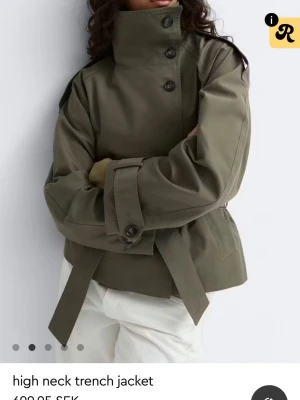 Olivgrön trenchcoat med hög krage - Stilren olivgrön trenchcoat med hög krage och knäppning framtill. Jackan har bälte i midjan, breda ärmar med spänne vid handlederna och axelklaffar. Tillverkad i ett kraftigt tyg som ger en cool och modern siluett.