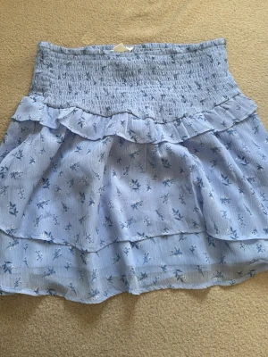 Blommig volangkjol från Gina Tricot - Söt ljusblå kortkjol från Gina Tricot (Gina young i st 170)  med blommigt mönster och volangdetaljer. Kjolen har smock i midjan för skön passform och är gjord i ett lätt, luftigt material som passar perfekt till varma dagar.