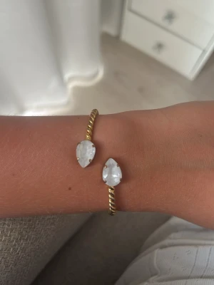 Caroline Svedblom Armband - Snyggt armband från Caroline Svedbom i guldtonad metall med tvinnad design. Armbandet är öppet och pryds av två droppformade vita kristaller i varje ände som ger en lyxig känsla. Perfekt accessoar för att lyfta din outfit.