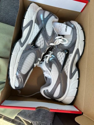 Nike vita och grå sneakers - Nike sneakers i vitt och grått med meshpaneler och detaljer i silver. Skorna har en chunky sula och klassisk snörning. Swoosh-logga på sidan och ovandel i syntet och mesh för bra andningsförmåga.