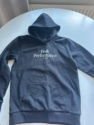 Svart hoodie Peak Performance - Snygg svart hoodie från Peak Performance med vit broderad logga och text 'Est. 1986' på bröstet. Klassisk känguruficka framtill och ribbade muddar vid ärmslut och nederkant. Storlek 160 barn men funkar på vuxen också