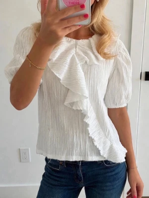 Vit blus med volangkrage - Säljer en vit blus i krinklat tyg med stor volangkrage och puffiga korta ärmar. Blusen har en avslappnad passform och ger en trendig och romantisk vibe. Perfekt att styla med jeans eller kjol för en cool look.