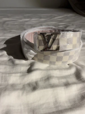 Vit och grå rutigt bälte  - Snyggt bälte i vitt och grått med rutigt mönster och en stor silvrig spänne framtill. Perfekt accessoar för att lyfta din outfit med en exklusiv touch. Bältet har en klassisk design och passar till många olika stilar.