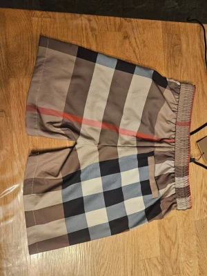 Burrberry bad shorts. Äkta. Pris kan diskuteras. Helt oanvända - Snygga shorts med stort rutmönster i beige, brunt, blått, vitt och en röd detalj. Shortsen har elastisk midja med snörning och sidofickor. Perfekta för sommaren och ger en trendig look.