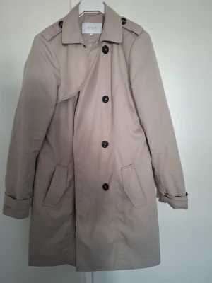 Beige trenchcoat från Vila - Stilren beige trenchcoat från Vila med klassisk dubbelknäppning, axelklaffar och två snedställda fickor. Jackan har ett bälte i midjan och svarta knappar som kontrast. Perfekt för dig som vill ha en tidlös och trendig look.
