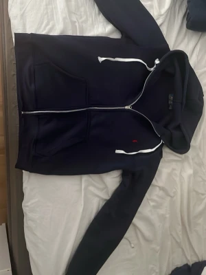 Mörkblå hoodie med dragkedja ralph lauren - Snygg mörkblå hoodie från Polo Ralph Lauren med vit dragkedja, vita snören i huvan och klassisk röd logga på bröstet. Hoodien har kängurufickor framtill och är tillverkad i mjukt material som känns skönt mot huden. Perfekt för en avslappnad och stilren look.