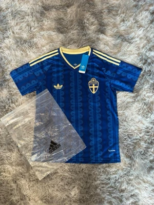 Svergie Vm tröja - Snygg blå fotbollströja från Adidas med Sveriges landslagsemblem på bröstet och gula detaljer. Tröjan har klassiska Adidas-ränder på axlarna, v-ringad krage och ett unikt mönster i tyget. Perfekt för match eller träning.