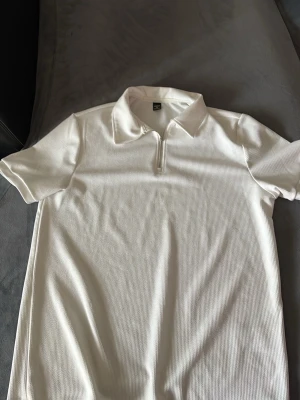 Vit kortärmad skjorta med half zip - Säljer en stilren vit skjorta med korta ärmar och krage. Skjortan har en half zip-dragkedja framtill och är tillverkad i ett lätt, strukturerat material som känns skönt mot huden. Perfekt för dig som gillar en clean och enkel look.