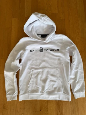 Vit hoodie från Sail Racing - Vit hoodie från Sail Racing med svart logga på bröstet. Klassisk känguruficka framtill och mjuk insida. Snygg och enkel design med huva och ribbade muddar. Perfekt för dig som gillar stilrena plagg och vill ha något bekvämt till vardags.