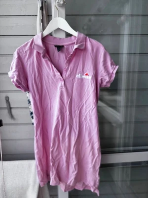 Rosa pikétopp från Ellesse - Säljer en ljusrosa pikétopp från Ellesse med klassisk krage och korta ärmar. Toppen har knappar framtill och broderad logga på bröstet. Perfekt för en sportig och avslappnad stil.