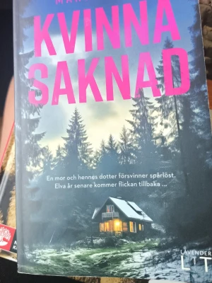 Kvinna saknad - Spännande skönlitterär bok med titeln 'Kvinna saknad'. Omslaget visar en mystisk stuga i en mörk skog med snö på marken och lysande fönster. Texten är tryckt i starkt rosa mot en blågrå bakgrund. Perfekt för dig som gillar thrillers och mysterier.