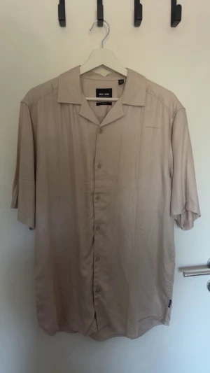 Beige kortärmad skjorta Only & Sons - Stilren beige skjorta från Only & Sons med korta ärmar och klassisk krage. Skjortan har knappar framtill och en avslappnad passform, perfekt för varma dagar. Tillverkad i ett mjukt och svalt material. Skjortan är helt ny, har tyvärr tagit bort prislappen men den är enbart testad vid köp. 