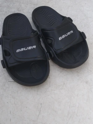 Svarta sandaler från Bauer storlek 35 - Säljer ett par svarta sandaler från Bauer med justerbara kardborreband och präglad sula för extra grepp. Tillverkade i slitstark plast, perfekta för chill dagar eller efter träning. Klassisk sportig look med Bauer-logga på ovandelen. Pris kan diskuteras
