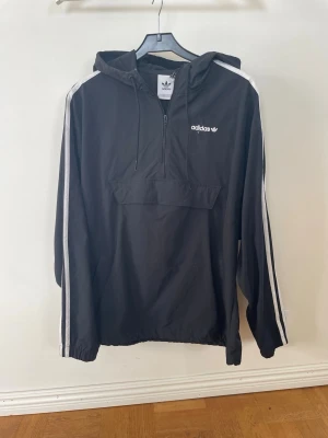 Svart vindjacka med huva från Adidas - Svart vindjacka från Adidas med vit logga på bröstet och klassiska vita ränder längs ärmarna. Jackan har huva med dragsko, halv dragkedja framtill och en stor magficka. Tillverkad i lätt och vindtätt material, perfekt för blåsiga dagar.