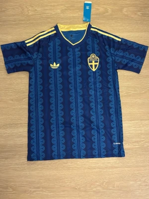 Blå Sverige fotbollströja Adidas - Snygg blå fotbollströja från Adidas med Sveriges landslagsemblem på bröstet och gula detaljer. Tröjan har ett retroinspirerat mönster, gula ränder på axlarna och V-ringad krage. Perfekt för match eller träning.