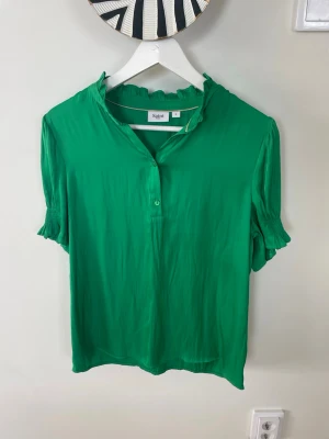 Grön blus från Saint Tropez - Snygg grön blus från Saint Tropez med korta ärmar och volangkant vid halsen. Blusen har knäppning framtill och lätt glansigt material som ger en fräsch look. Perfekt till jeans eller kjol för en trendig stil.