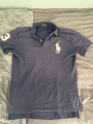 Mörkblå pikétröja från Polo Ralph Lauren - Klassisk mörkblå pikétröja från Polo Ralph Lauren med vit broderad logga på bröstet och siffran 3 på ärmen. Tröjan har krage, knappar vid halsen och korta ärmar. Perfekt för en stilren och sportig look.
