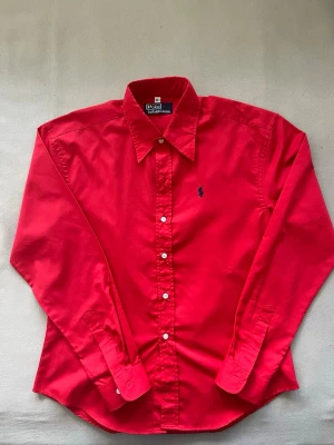 Ralph lauren skjorta - Snygg röd skjorta från Polo Ralph Lauren med klassisk krage och knäppning framtill. Skjortan har långa ärmar och den ikoniska broderade loggan på bröstet. Perfekt för dig som vill sticka ut med färg och stil.