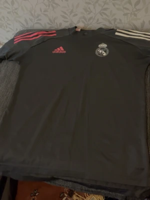 Grå Real Madrid träningströja Adidas - Snygg grå träningströja från Adidas med Real Madrid-logga på bröstet. Tröjan har rosa Adidas-logga och rosa samt vita ränder på axlarna. Perfekt för fotbollsträning eller chill. Tillverkad i ett lätt och funktionellt material.