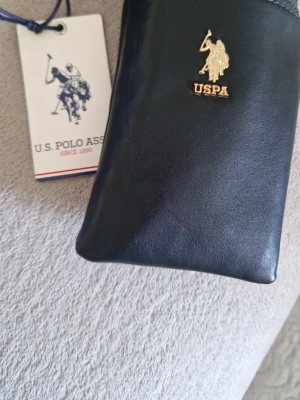 Svart miniväska från U.S. Polo Assn. - Snygg svart miniväska från U.S. Polo Assn. i slätt konstläder med guldfärgad logga framtill. Perfekt för det viktigaste och enkel att matcha till olika outfits. Klassisk och stilren design med exklusiv känsla.
