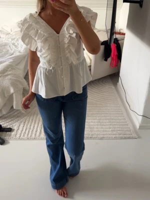 Vit blus med volanger och knappar - Supersöt vit blus med volanger längs axlar och bröst, v-ringning och knappar framtill. Blusen har en lätt utsvängd passform och korta ärmar. Perfekt att styla med jeans eller kjol för en trendig look.