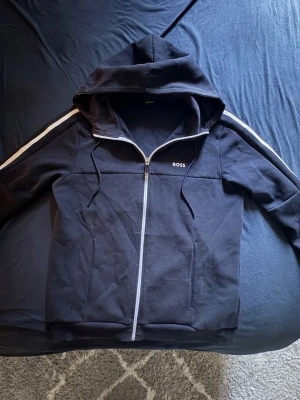 Svart hoodie med dragkedja från BOSS - Snygg svart hoodie från BOSS med vit dragkedja och vita ränder längs ärmarna. Klassisk huva med dragsko och två fickor framtill. Tillverkad i mjukt material som känns skönt mot huden. Perfekt för en avslappnad och stilren look.(((((     PASSAR ÄVEN M   ))))