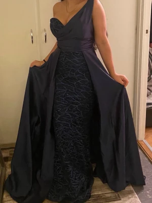 Marinblå långklänning oneshoulder - Snygg marinblå långklänning med oneshoulder-design och elegant omlott över bysten. Kjolen har ett glittrigt mönster och ett lager av släpande tyg som ger extra volym. Perfekt för bal eller festliga tillfällen. Passar xs / s