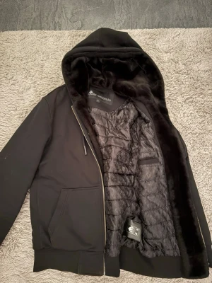 Svart bunnyjacka med huva - Säljer en svart bunnyjacka från Moose Knuckles med mjukt foder och stor huva. Jackan har dragkedja, fickor framtill och en patch med metallogo på ärmen. Insidan är fodrad med fluffigt material för extra värme och komfort.