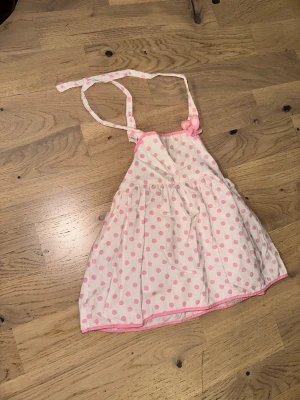 Vit och rosa prickig kort klänning - Supersöt kort klänning i vitt med rosa prickar och tunna axelband som knyts i nacken. Klänningen har en lätt utställd kjol och rosa kantdetaljer. Perfekt för varma dagar och ger en lekfull vibe.Vit och rosa prickig kort klänning