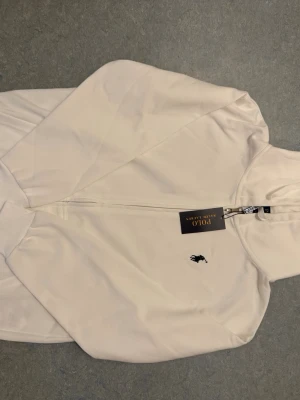 Vit hoodie från Polo Ralph Lauren - Snygg vit hoodie från Polo Ralph Lauren med dragkedja framtill och klassisk logga broderad på bröstet. Modellen är custom fit och har ribbade muddar samt huva. Perfekt för en clean och stilren look. Storlek L men passar även M